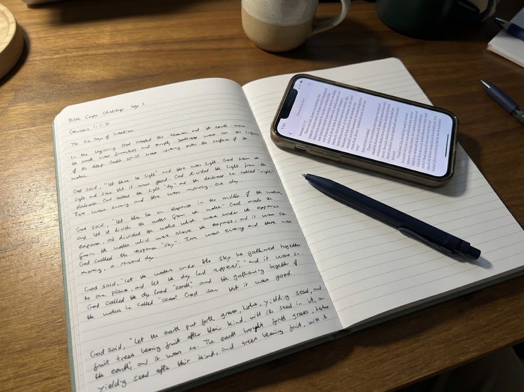 Day 1 of the Bible Copy Challenge copied into the journal, with the passage open on a phone